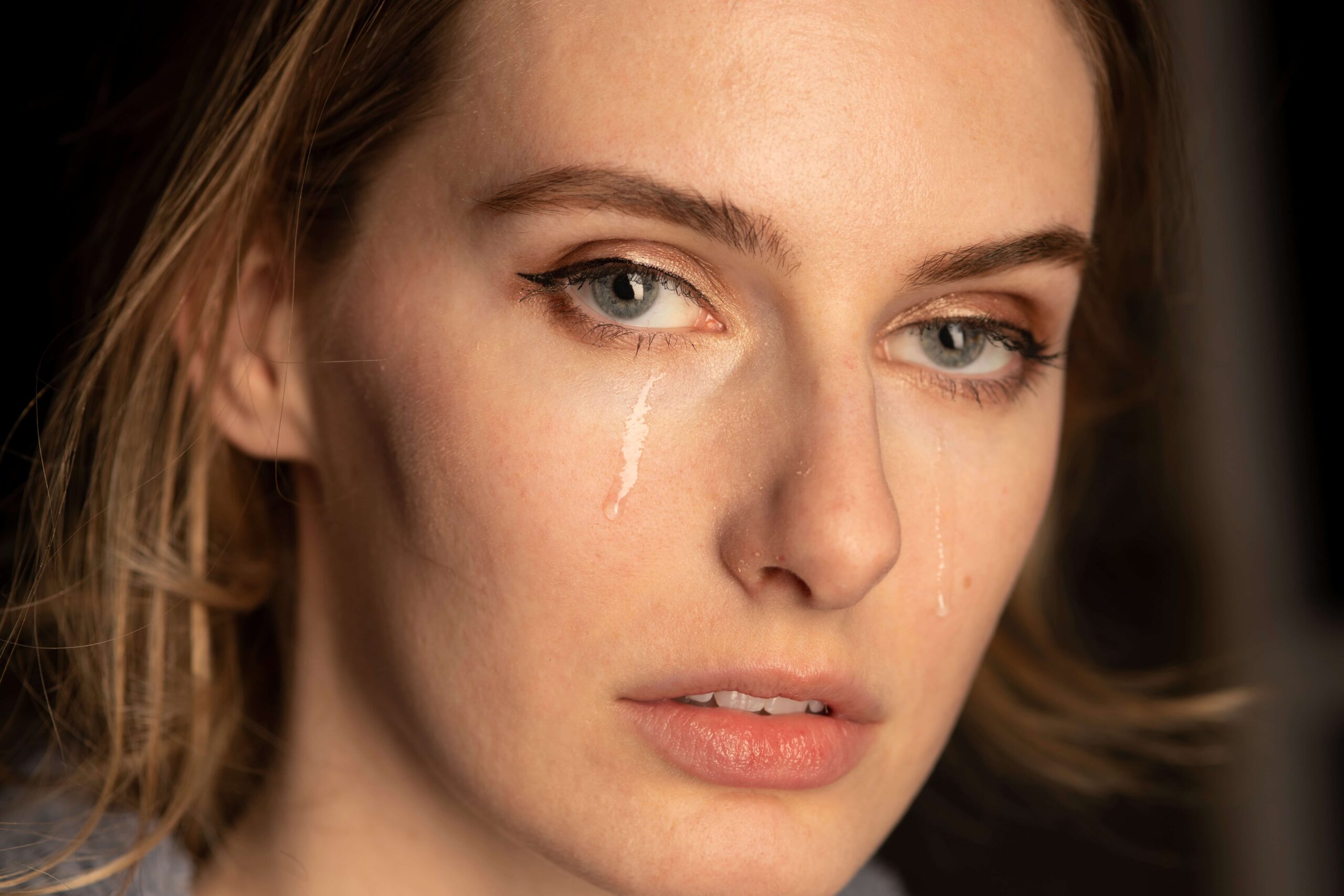 Nahaufnahme eines Gesichts mit heller Haut und blauen Augen. Zwei Tränen laufen jeweils von einem Auge die Wangen hinunter. Der Hintergrund ist dunkel und unscharf.