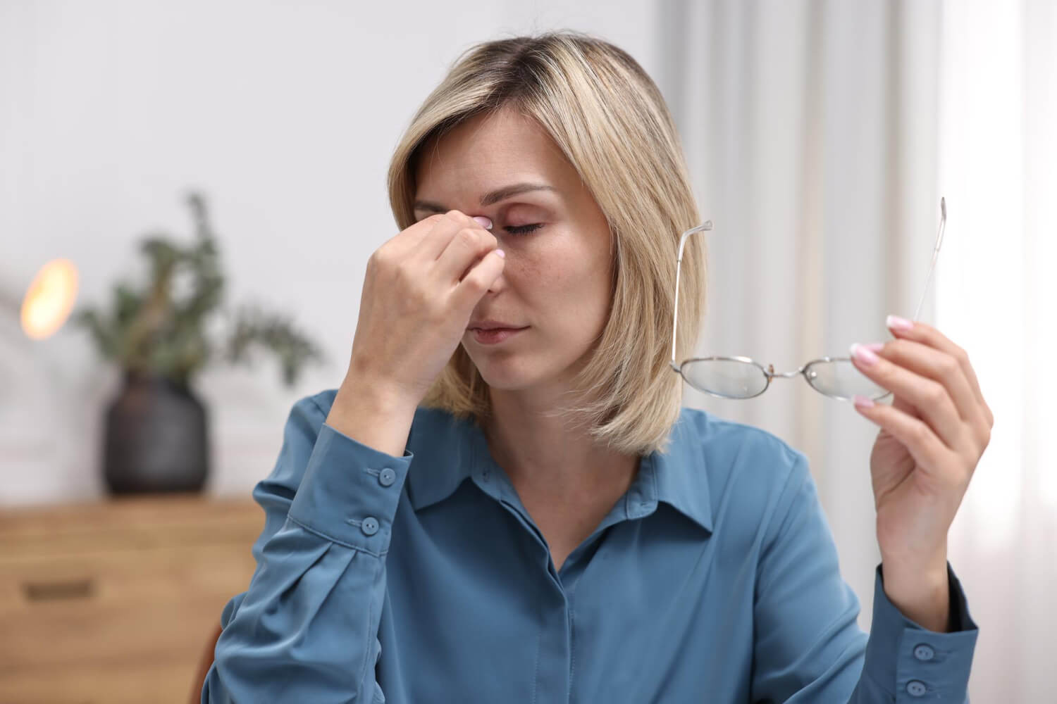 Eine Frau mit schulterlangem blondem Haar sitzt in einem Innenraum und hält eine Brille in der rechten Hand. Mit der linken Hand drückt sie sich die Nasenwurzel, die Augen sind geschlossen. Sie trägt eine blaue Bluse, im Hintergrund stehen Möbel und eine Pflanze.